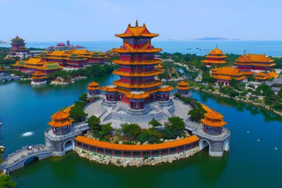 Colorful traditional buildings surround a calm blue lake, with the ocean in the background.